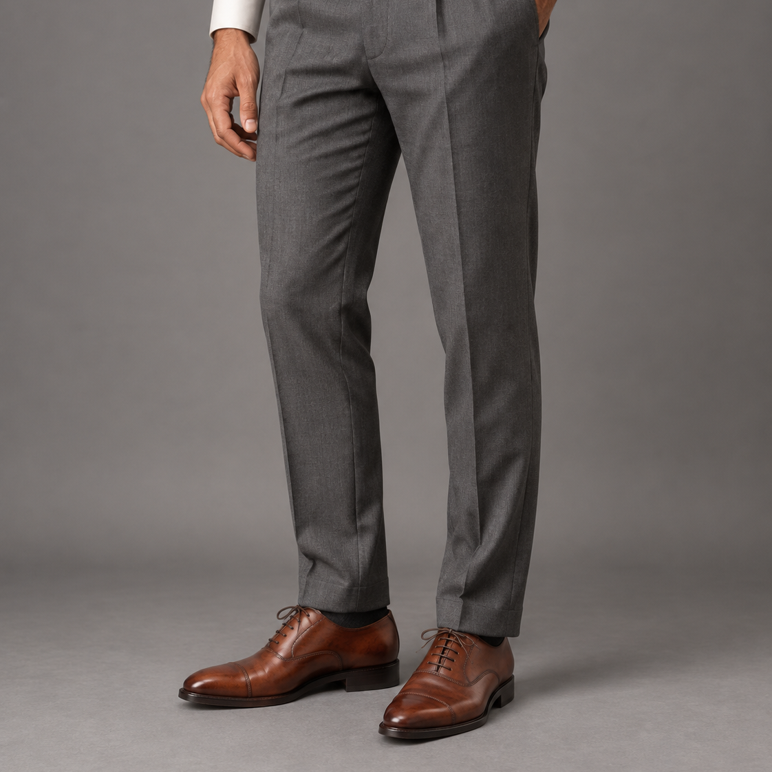 A person wearing gray pleated dress trousers with a brown belt, white dress shirt, and brown leather oxford shoes, shown from the waist down against a plain gray studio background.